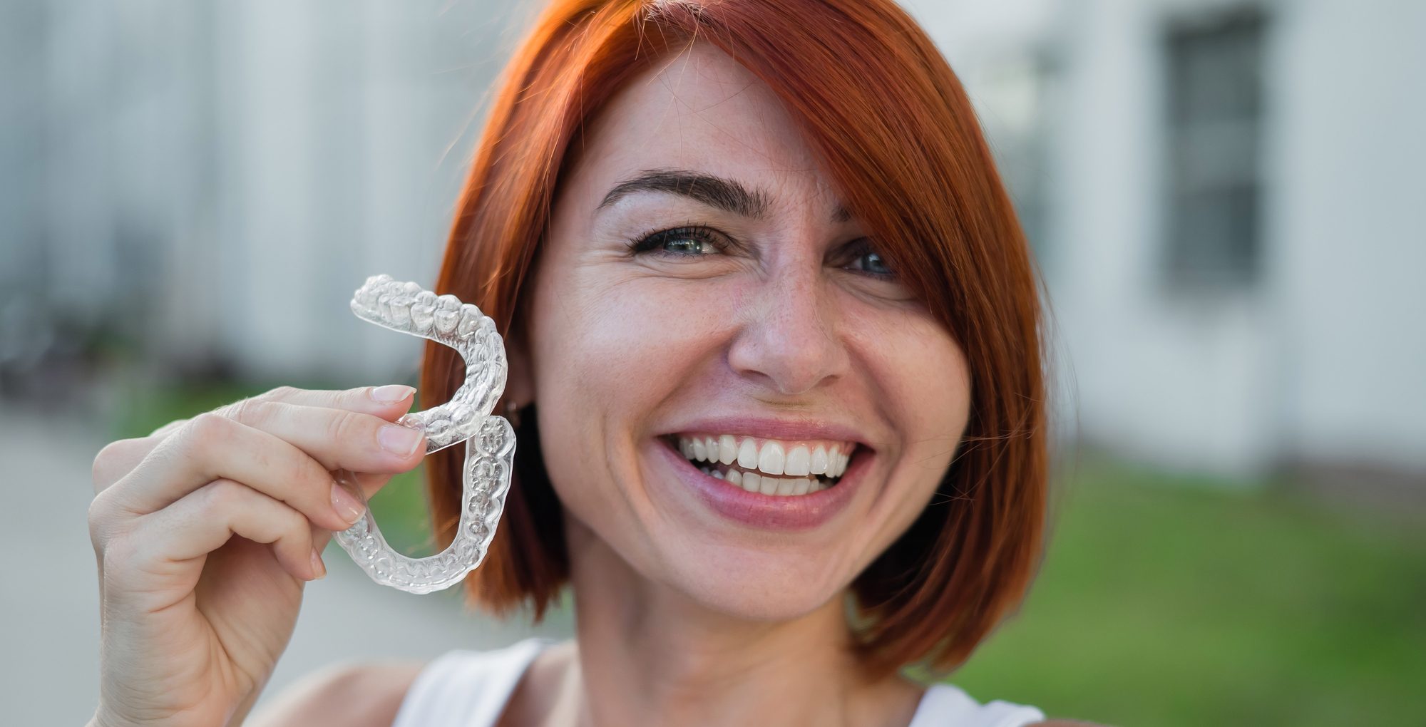 Red-haired Caucasian woman holding transparent mouthguards for bite correction outdoors. A girl with a beautiful snow-white smile uses silicone braces